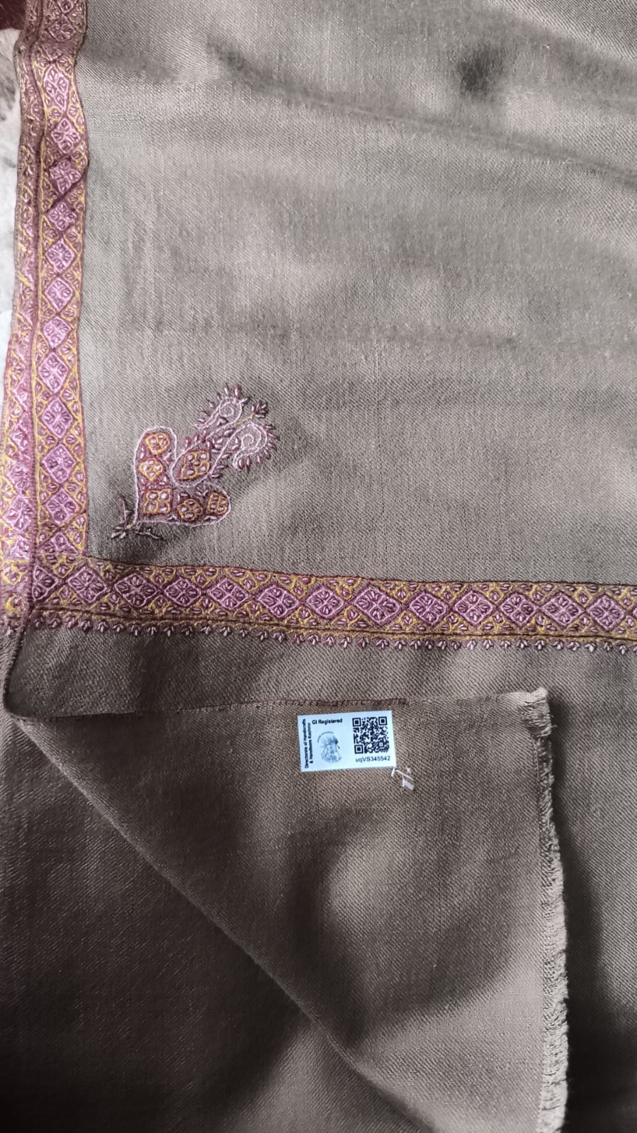 A close-up of a taupe brown shawl with a decorative border featuring pink, purple, and gold embroidery, and a small embroidered motif near the top left corner.