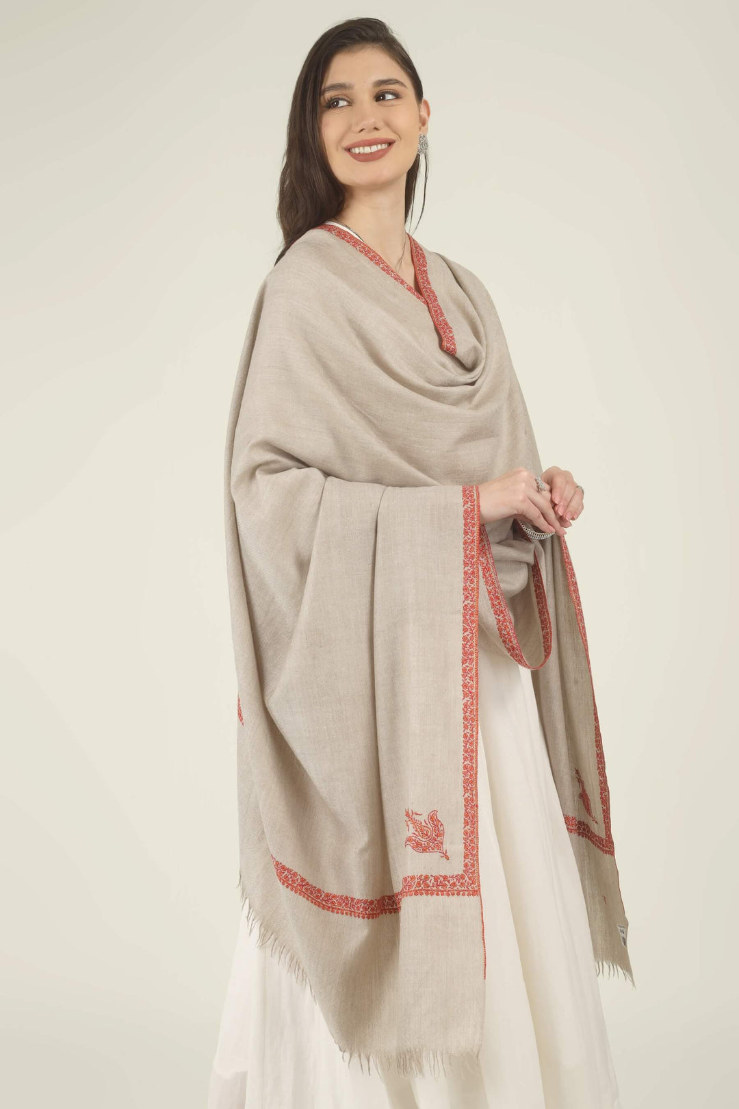 This Natural Khudrang GI Kashmiri Pure Pashmina Hashidaar Embroidered Shawl features a soft, undyed oatmeal-beige body framed by richly worked borders and paisley motifs in vivid red and warm orange