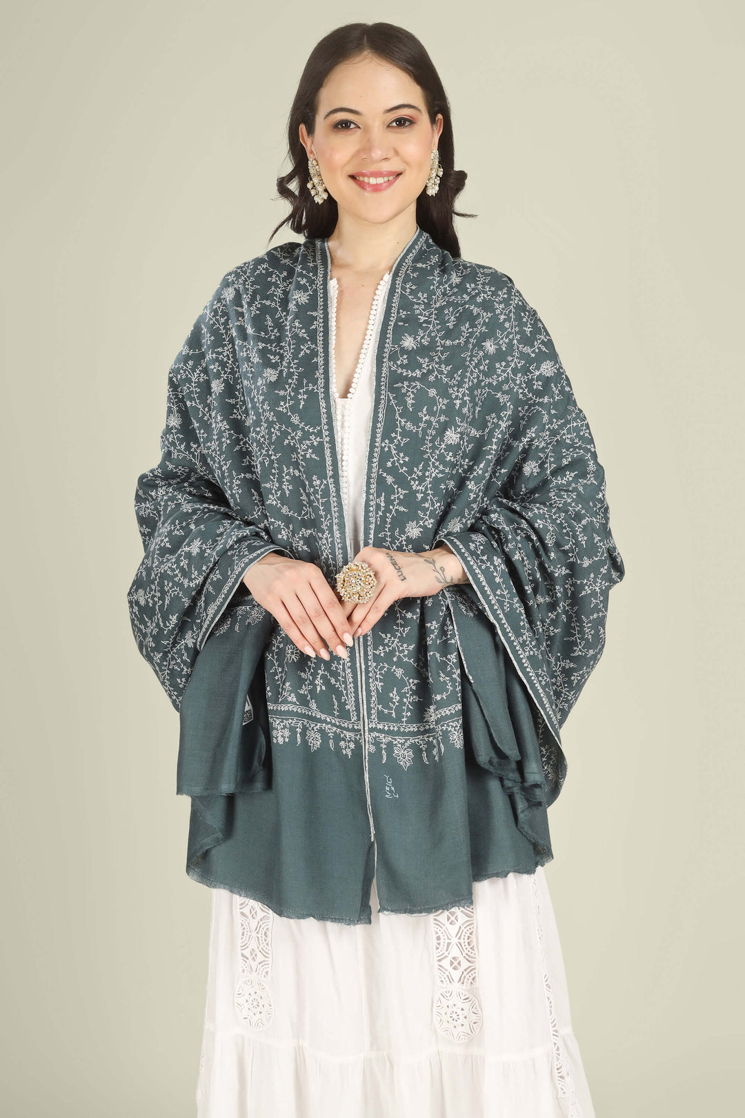 A woman modeling a dark teal-green shawl with white floral block prints and white border embroidery, worn over a white dress against a solid light beige background.