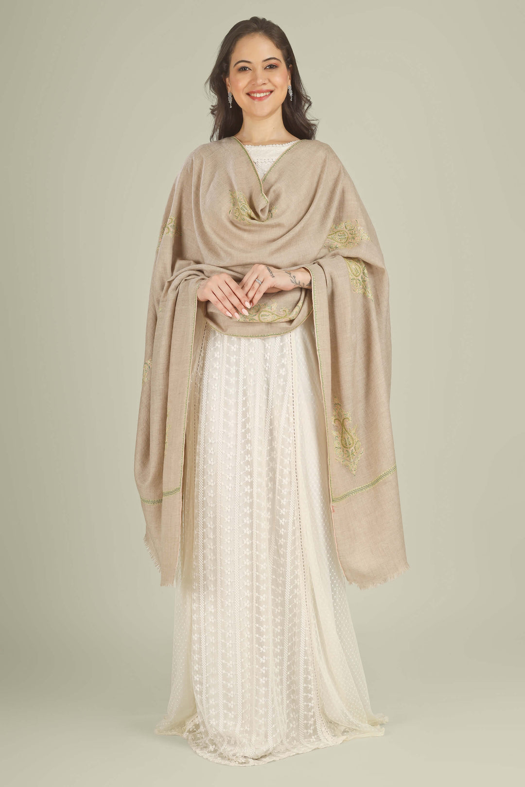 A woman modeling a draped beige shawl with gold embroidery over a white embroidered dress against a solid light beige studio background.