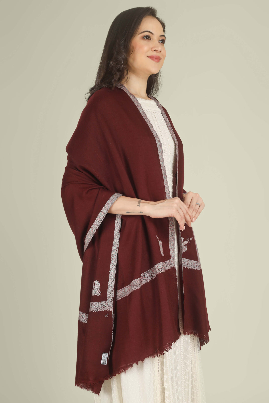 A woman wearing a deep maroon shawl with silver embroidered borders over a white dress, standing against a solid light beige background.