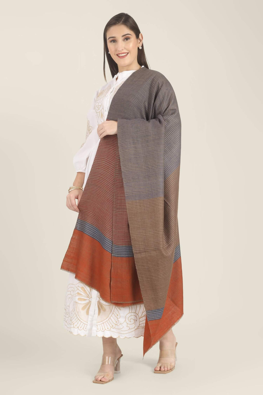 Model draped in a multicolour Pure Kashmiri Pashmina shawl with woven grey-blue, rust-red and taupe stripes and a wide plain rust border over a white embroidered dress.