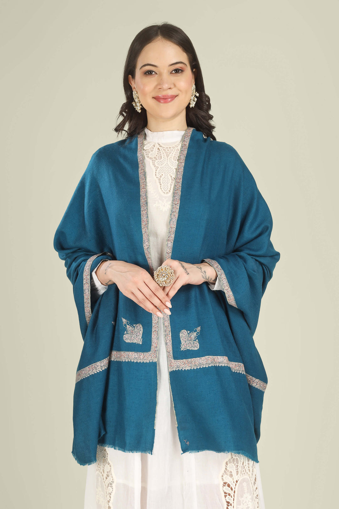 A woman models a teal blue shawl with silver embroidered details over a white lace-trimmed dress against a solid light beige background.