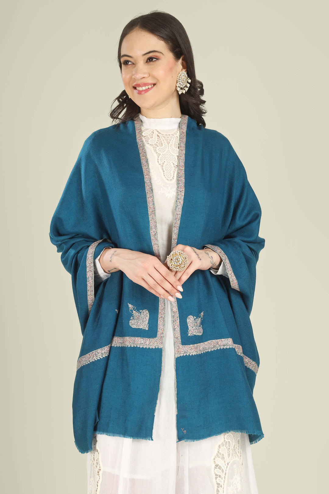 A woman wearing a teal blue shawl with silver embroidered borders over a white embroidered top, photographed against a plain light beige studio background.