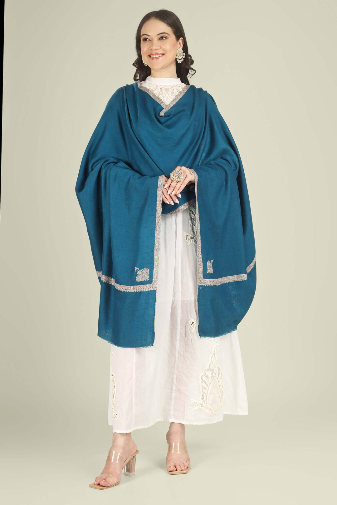 A woman modeling a teal blue shawl with silver embroidered borders over a white embroidered dress, standing against a solid light beige background.