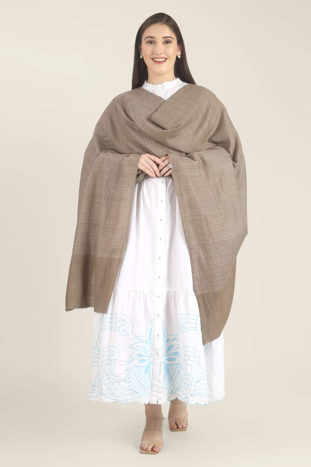 Model draped in a taupe Pure Kashmiri Pashmina shawl with fine woven pinstripes and a wide plain border over a white dress with light blue embroidery.