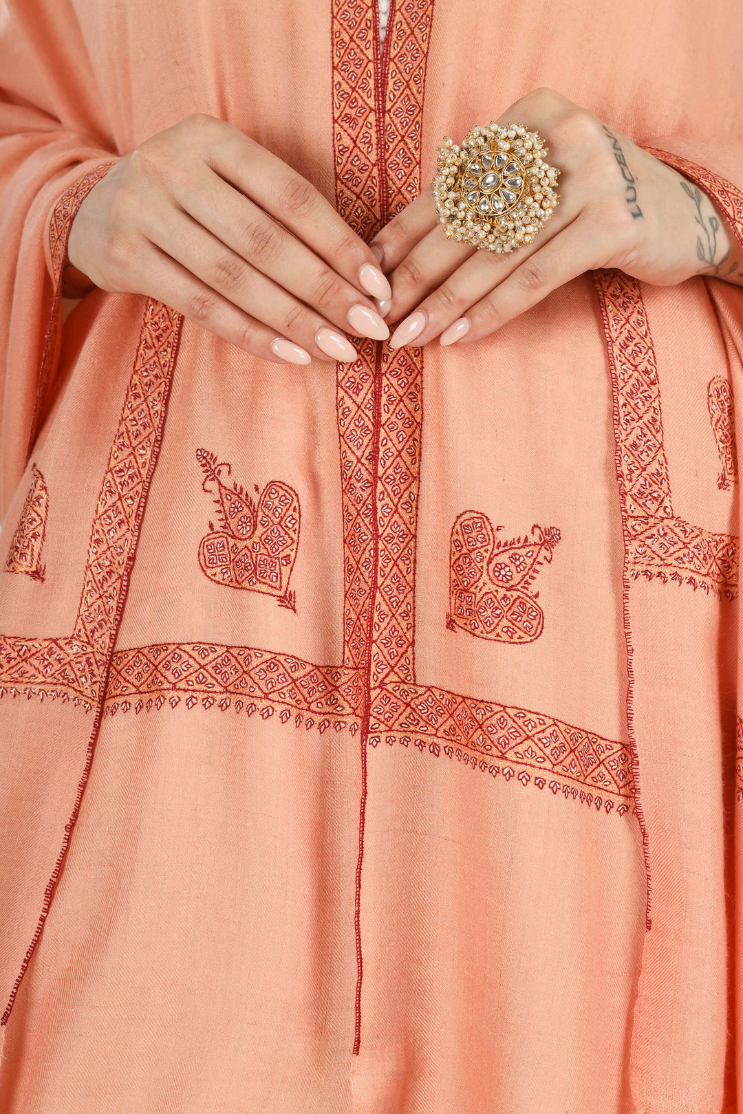 Close-up of a peach-colored embroidered kurta with red geometric borders and motifs, worn by a model whose hands are clasped in front. The model is also wearing a large gold ring.