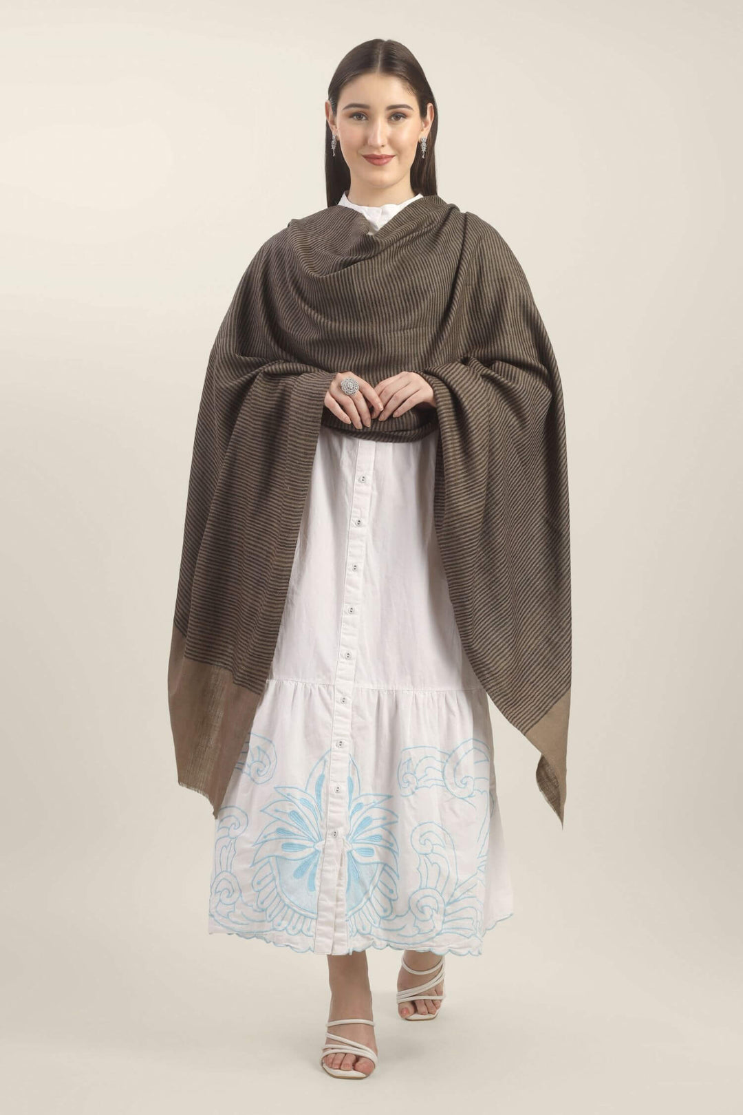 Model holding a taupe Pure Kashmiri Pashmina shawl with fine black woven stripes and a wide plain border over a white embroidered dress.